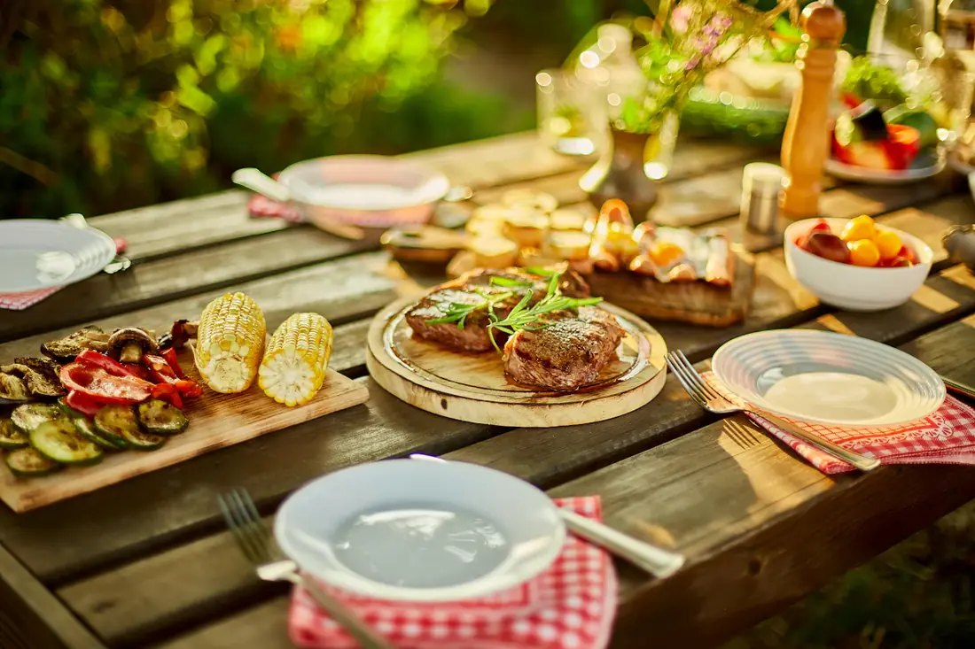 Grilled steaks, corn and vegetables on a wooden table set for an outdoor dinner